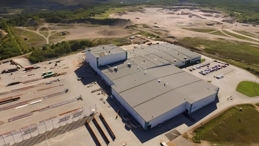 Aerial view of a grey industrial facility complex with a car park, storage yard, and surrounding cleared land