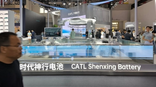 A trade show visitor walks past a battery exhibit booth with illuminated signage and a suspended car model